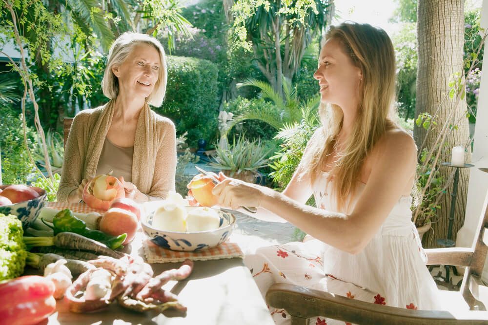 Wealthy mother and adult daughter peeling fruit at a table in a lush backyard garden