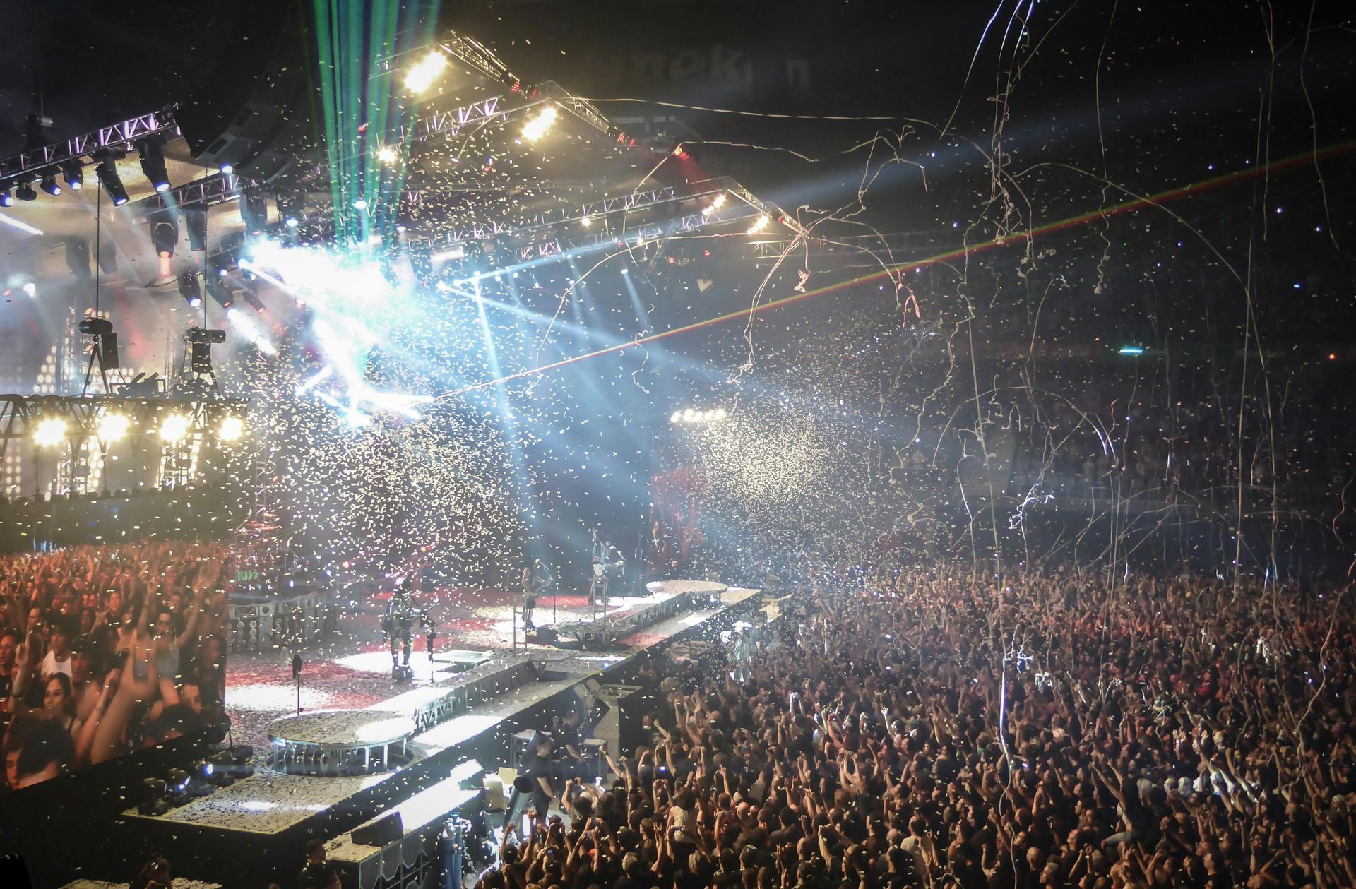 A packed concert arena filled with confetti, bright stage lights, and a cheering crowd reaching toward the performers.
