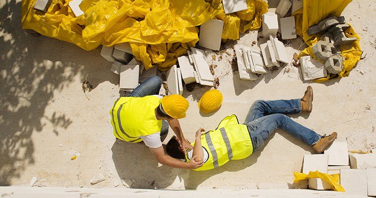 Aerial view of a builder in hard hat and high-vis tabar, helping a colleague who lies injured on the ground