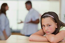 A sad-looking child at a table, resting her chin on her arms, as her parents argue behind her