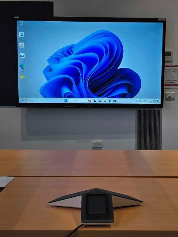 A conference room setup with a large display showing a Windows 11 background. A tabletop microphone is in the foreground.