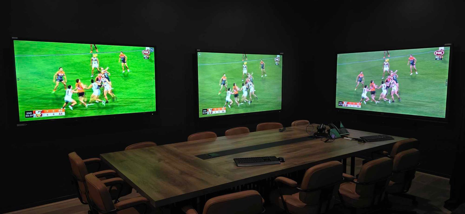 A conference room with three screens showing a sports game, a large wooden table, and chairs. The room is dark.