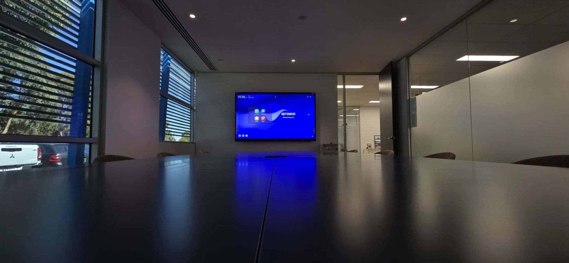 A modern conference room with a large blue TV screen. The room has large windows and a reflective dark table.