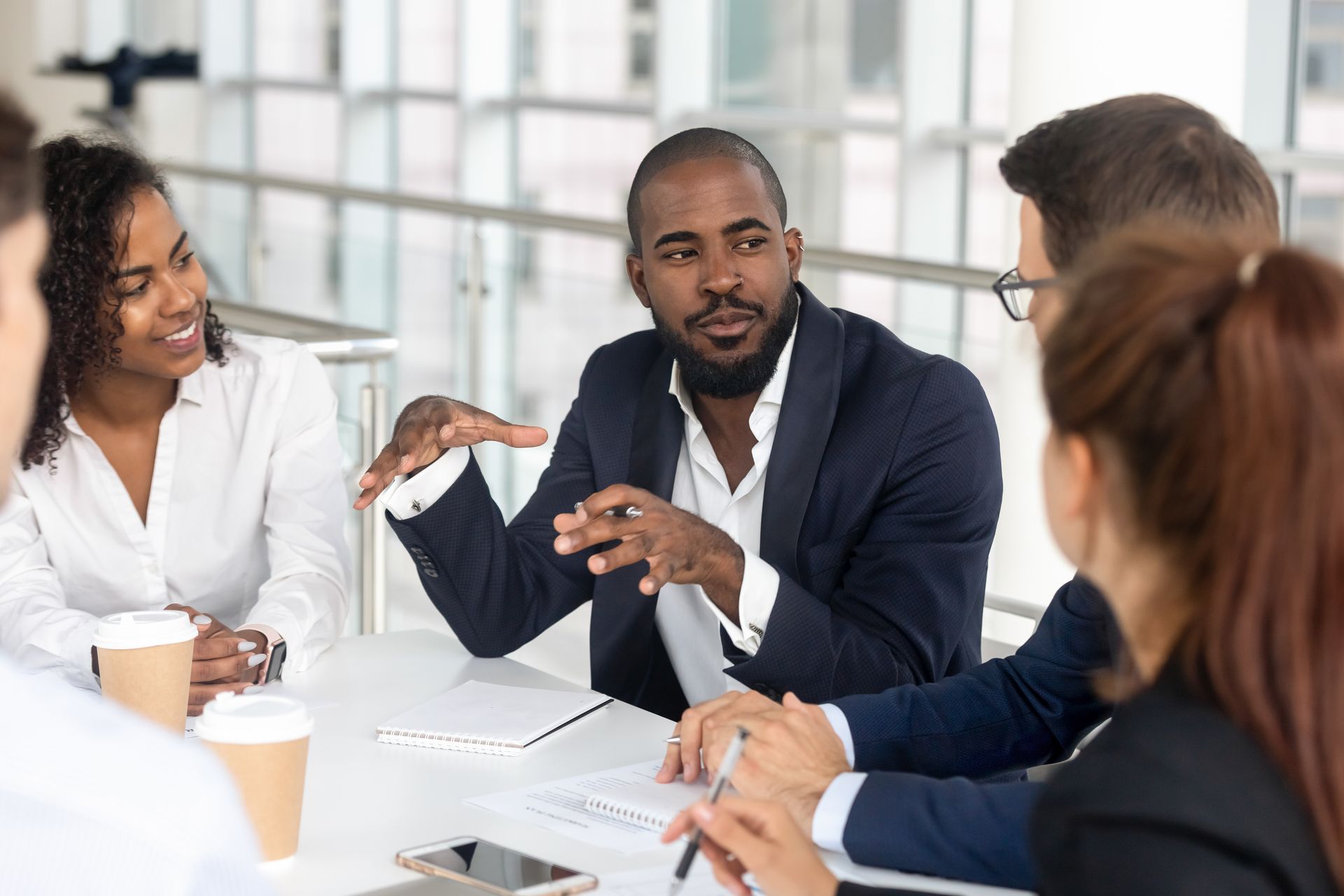 Group of people in business attire at a table, discussing a topic with documents and coffee present.