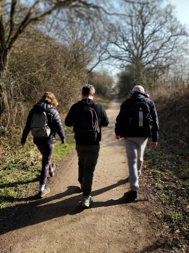 Three people walking on a dirt path in a wooded area, wearing jackets and backpacks.