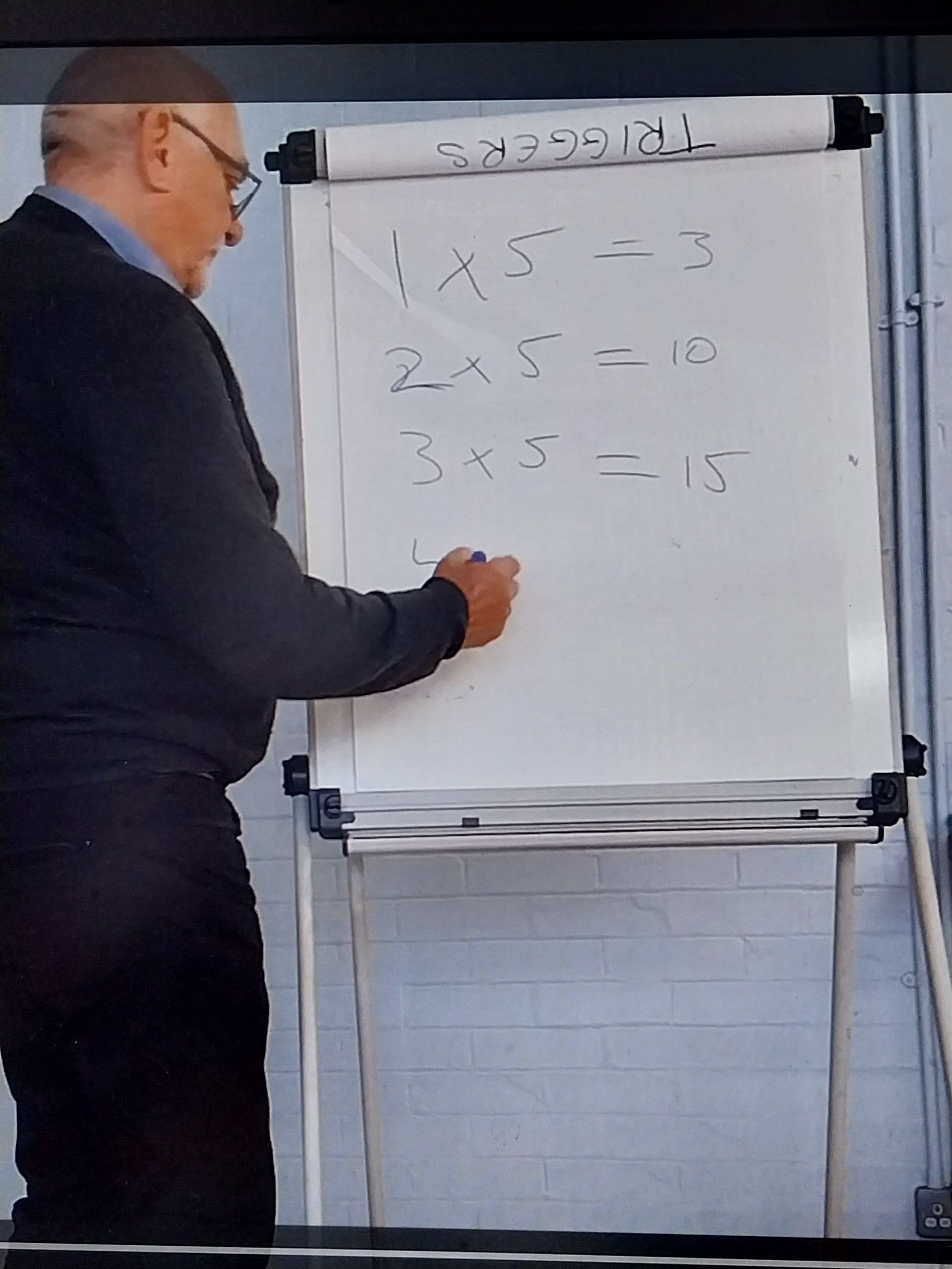 Man writing on a whiteboard with math equations: 1x5=5, 2x5=10, 3x5=15.