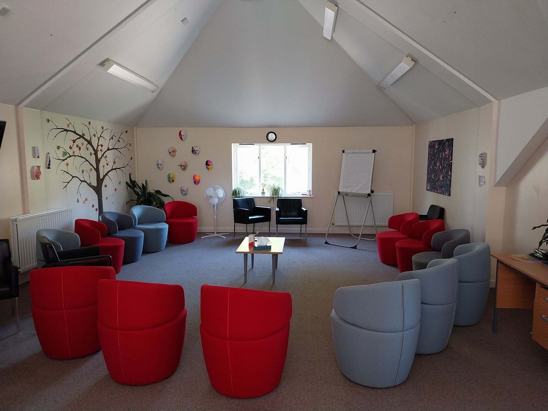 Meeting room with red and blue armchairs in a circle. Tree mural and whiteboard visible.