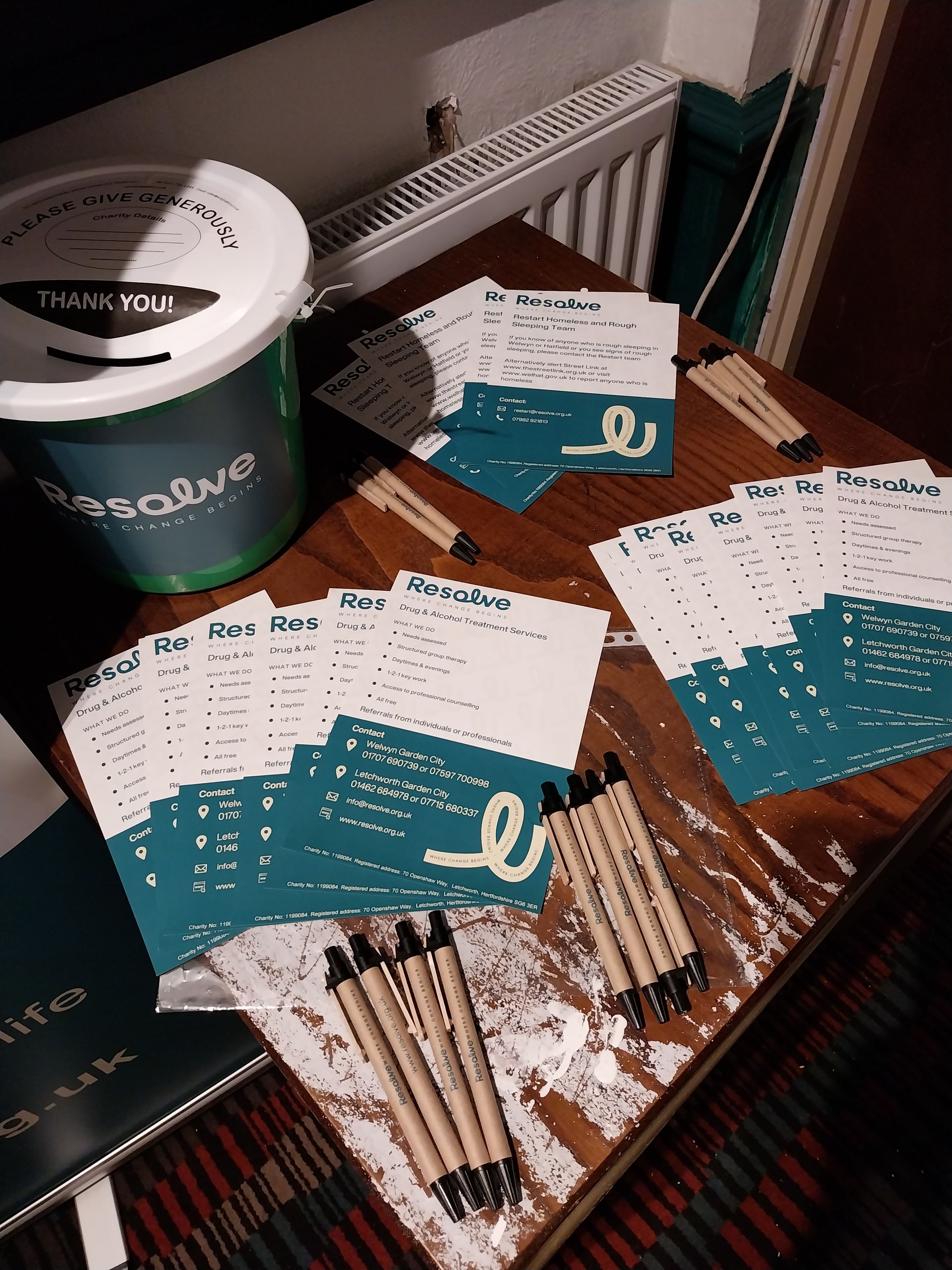 Wooden table with stack of teal-and-white cards, pens, and a donation bucket that reads
