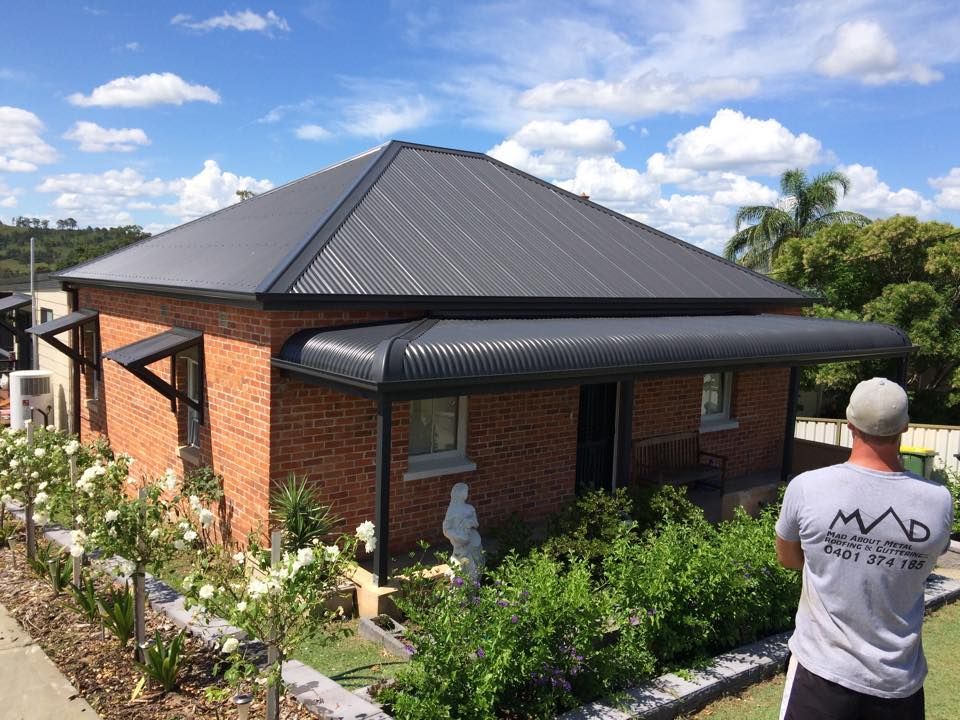 A Man Is Standing in Front of A Brick House with A Black Roof — Mad About Metal Roofing & Guttering PTY LTD in Newcastle, NSW