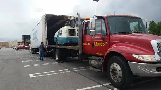 A red tow truck is carrying a trailer in a parking lot.