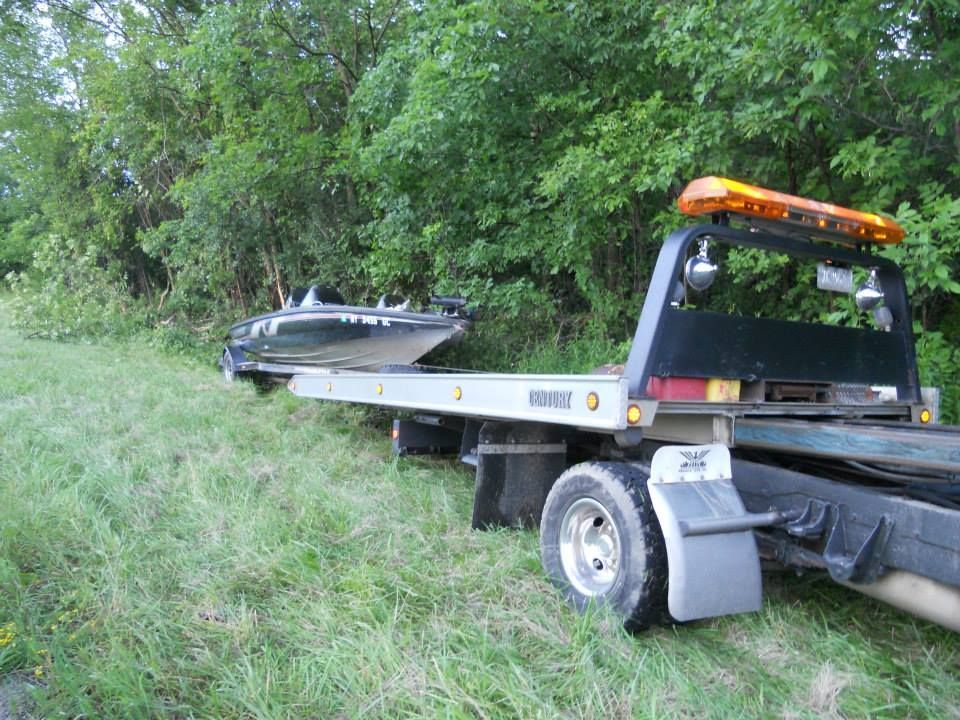 A tow truck with a boat on the back is parked in the grass.