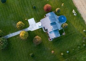 Aerial view of a house with a driveway and green lawn, surrounded by trees and a blue circular pool
