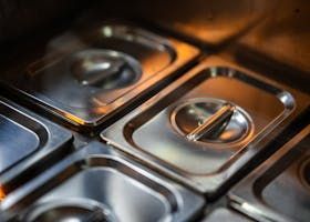 Close-up of stainless steel food containers with lids in a warm-lit kitchen counter tray.