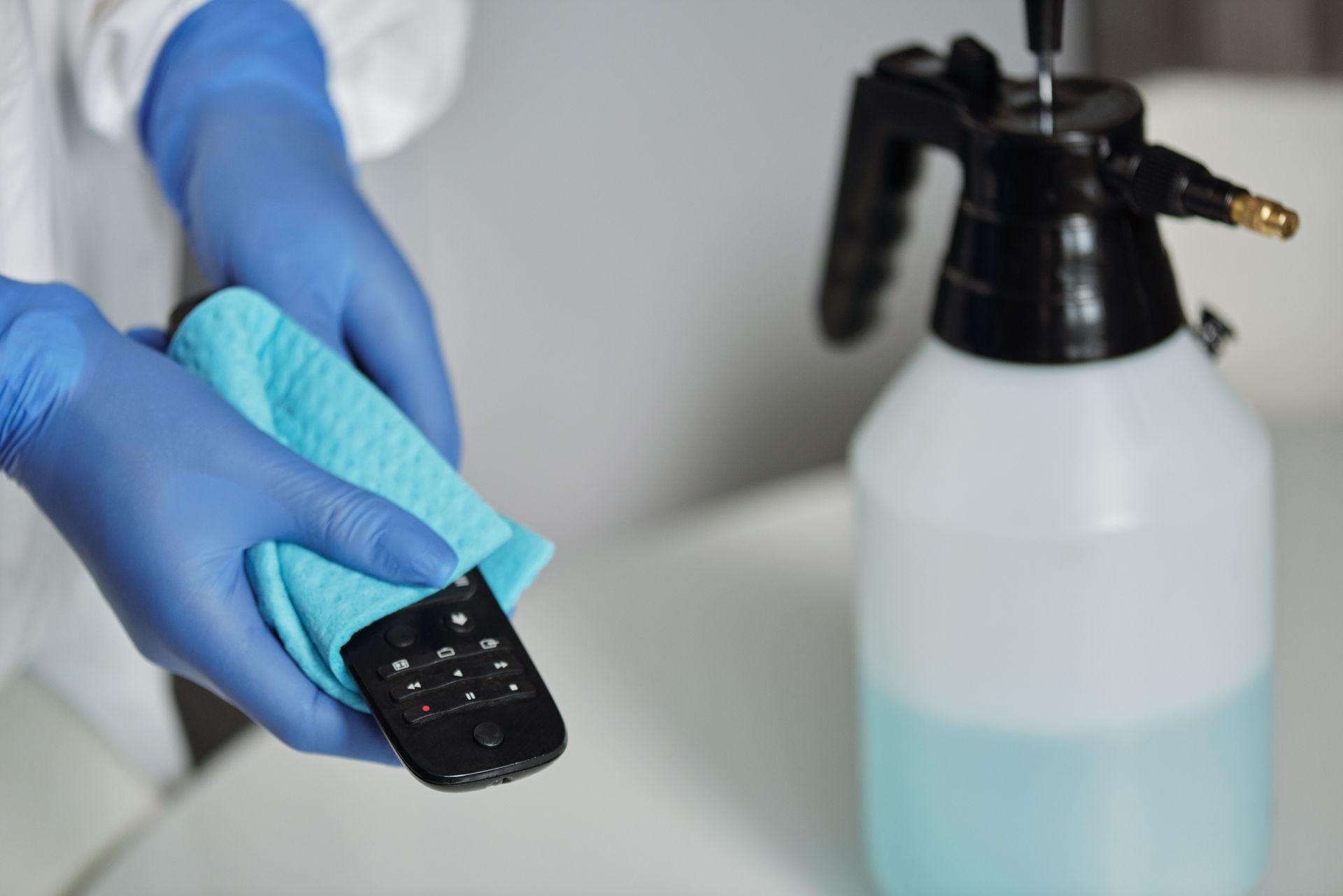 Gloved hands wiping a black remote with a blue cloth beside a spray bottle