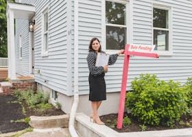 Woman standing by a red real estate sign outside a light gray house, holding papers and smiling