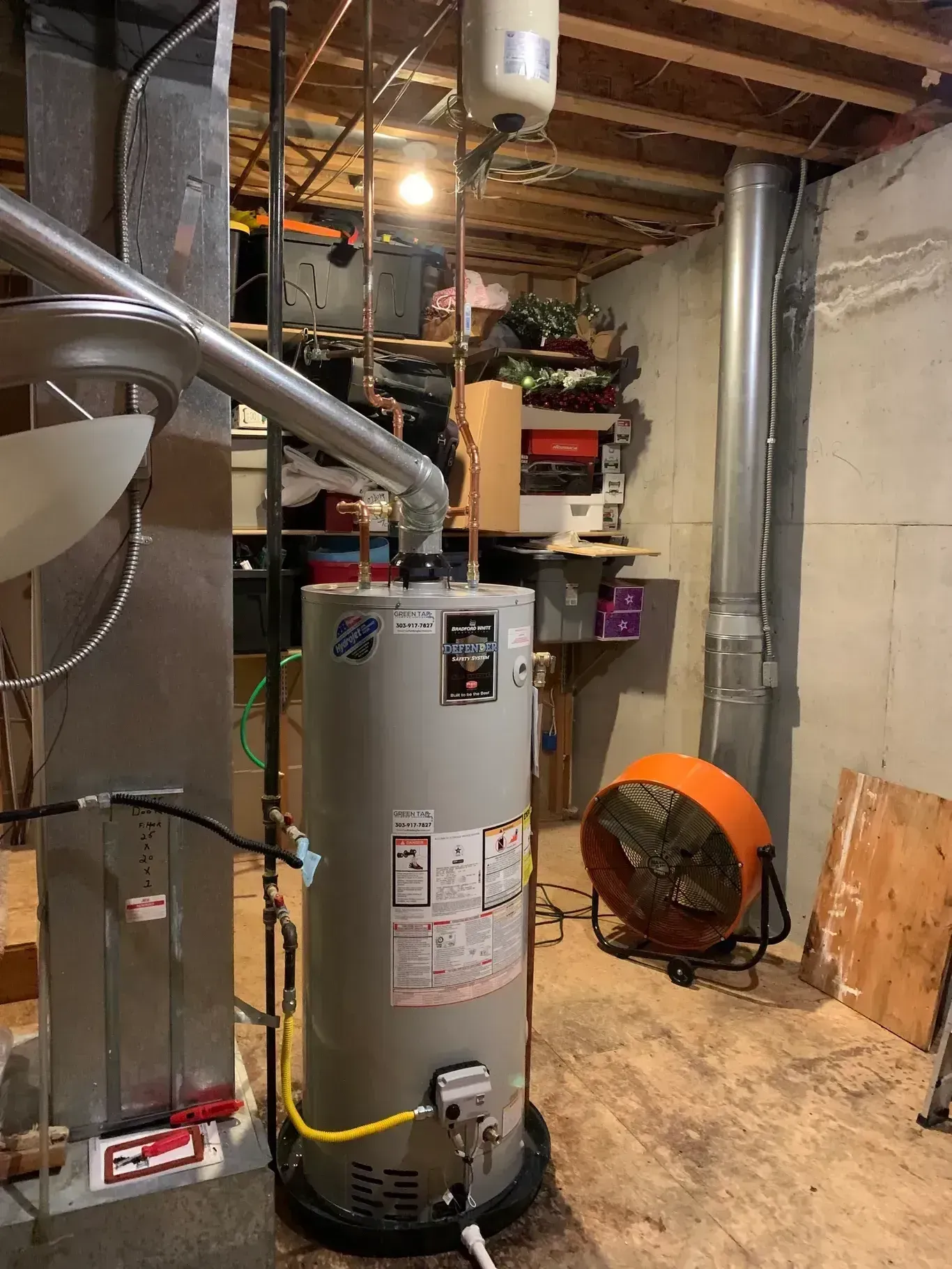 Water heater in a basement with metal vent pipes and an orange fan.