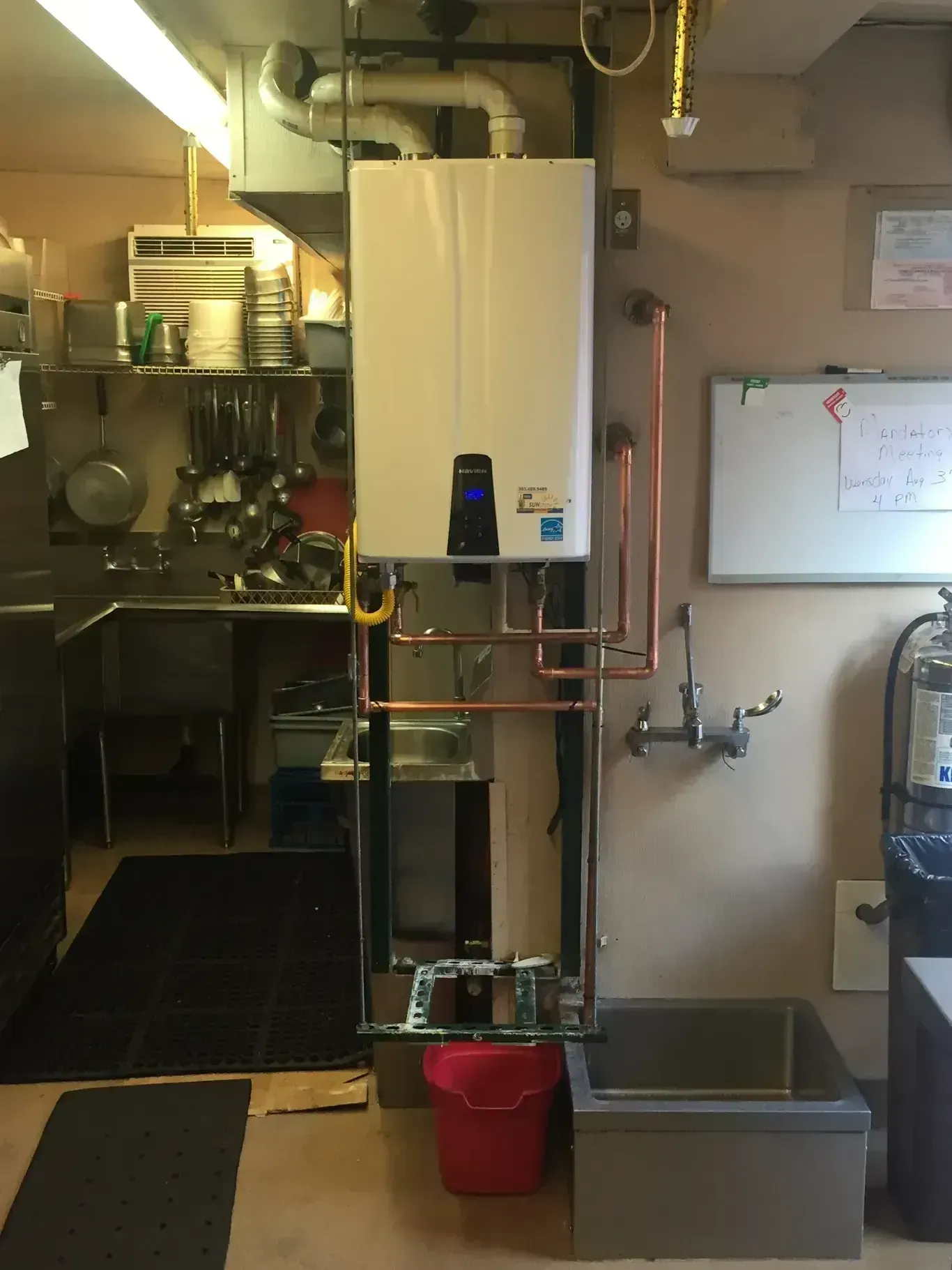 Wall-mounted water heater with copper pipes in a commercial kitchen next to a sink, with a red bucket below.