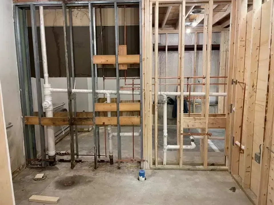 Interior view of a bathroom remodel in progress, with exposed pipes and wall studs.