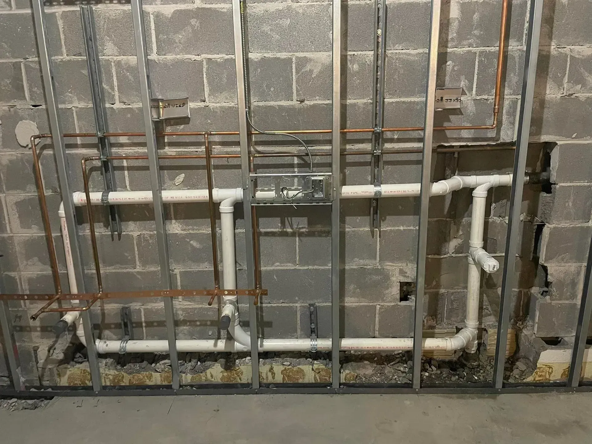 Metal studs, pipes, and electrical box against a concrete block wall, likely during construction.