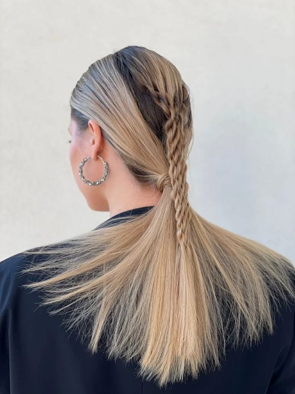 Capelli biondi raccolti in una coda bassa con una treccia morbida che scende lungo la schiena, visti di spalle contro una parete semplice.