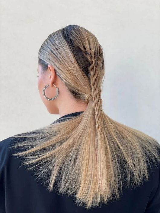 Capelli biondi raccolti in una coda bassa con una treccia morbida che scende lungo la schiena, visti di spalle contro una parete semplice.