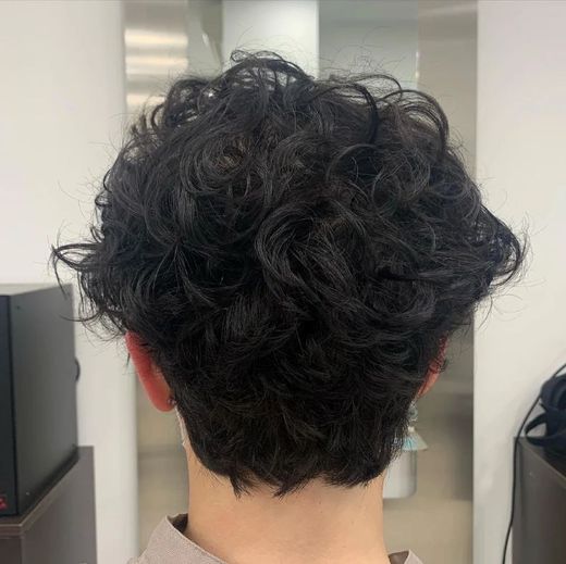 Vista posteriore di una persona con capelli scuri e ricci in un ambiente interno, con uno specchio e dei mobili alle sue spalle.
