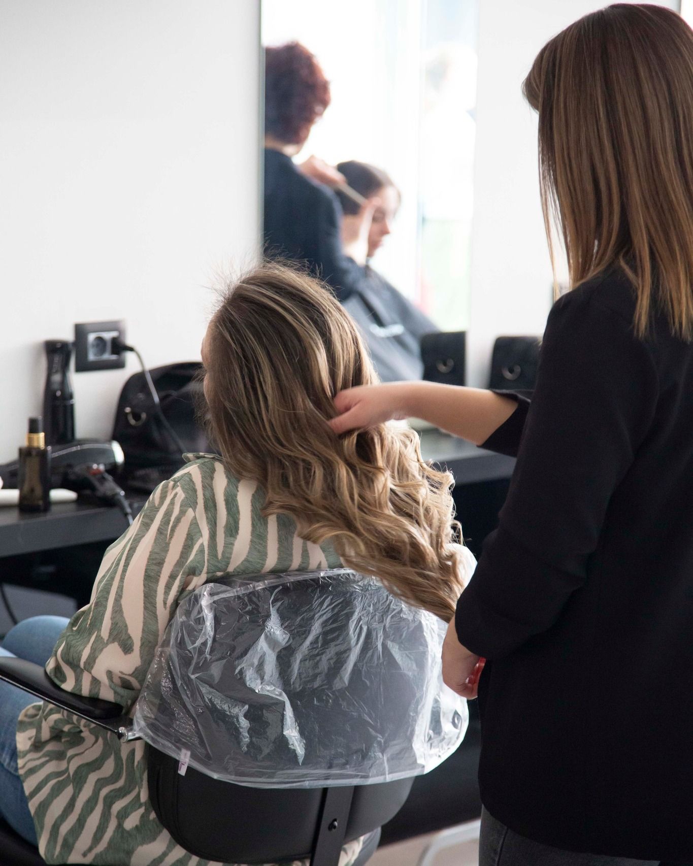 In un salone, una parrucchiera acconcia i capelli di una cliente, con specchi e poltrone da parrucchiere nere ben visibili.