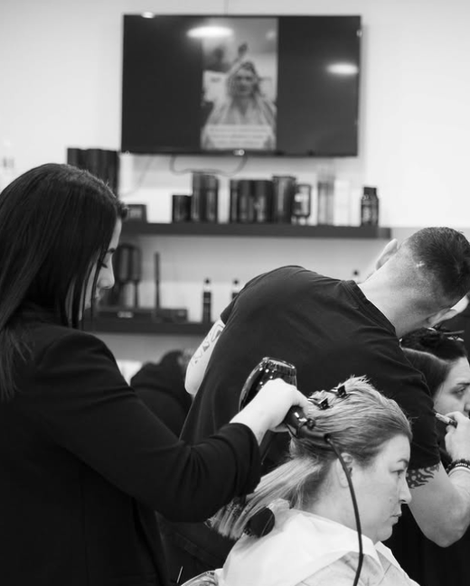 In un salone, alcuni parrucchieri lavorano sui clienti, con un televisore e degli scaffali sullo sfondo.