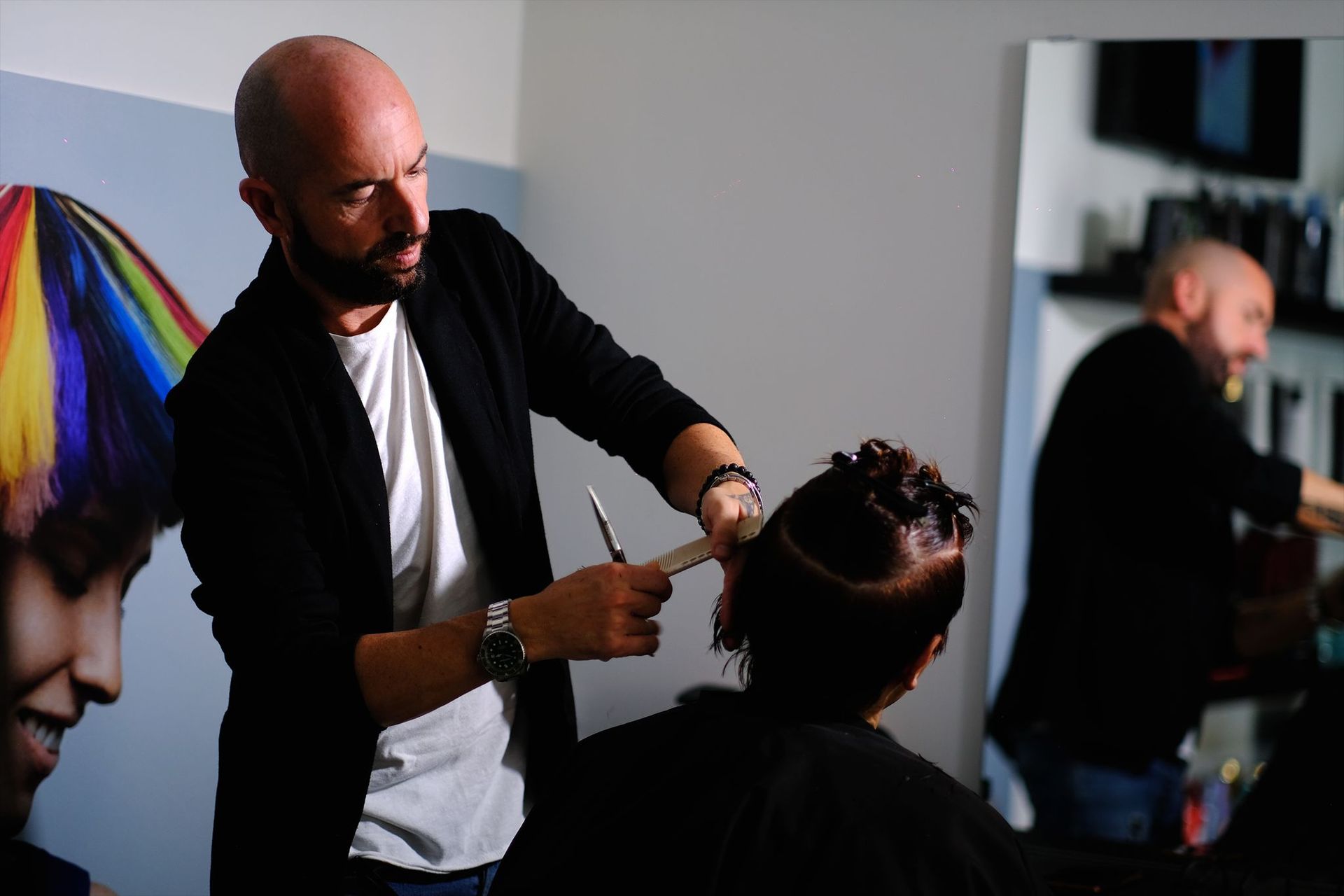 Un parrucchiere taglia i capelli a una cliente in un salone, con un poster colorato riflesso nello specchio.