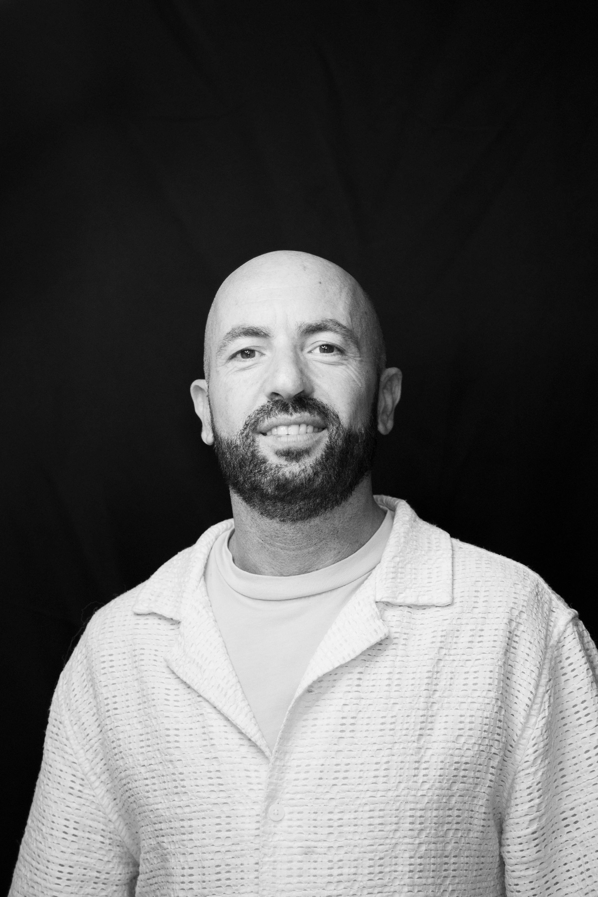 Ritratto in bianco e nero di un uomo calvo sorridente con la barba, su sfondo scuro.