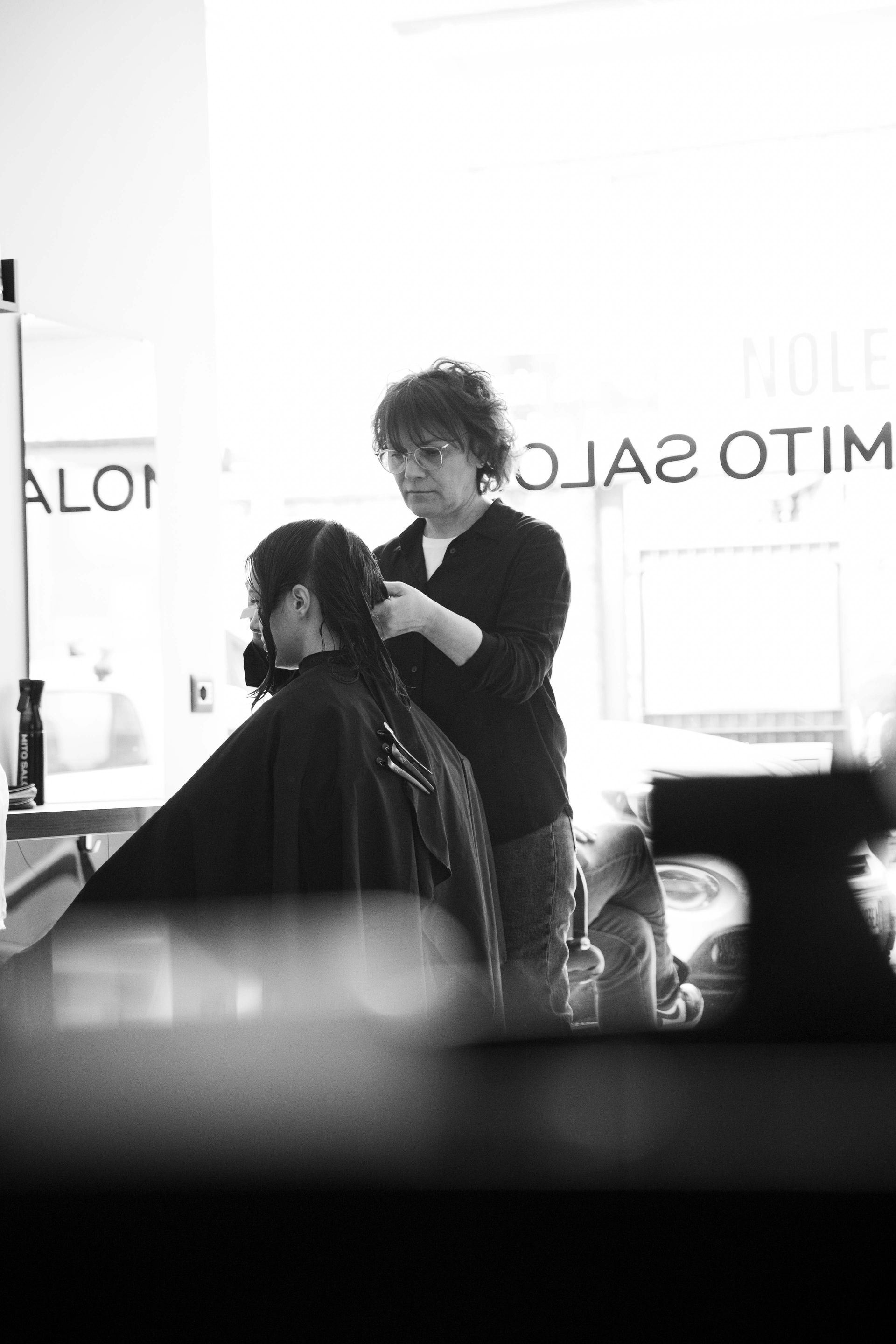 Un parrucchiere taglia i capelli scuri di una cliente in un salone, con specchi e finestre luminose alle sue spalle.