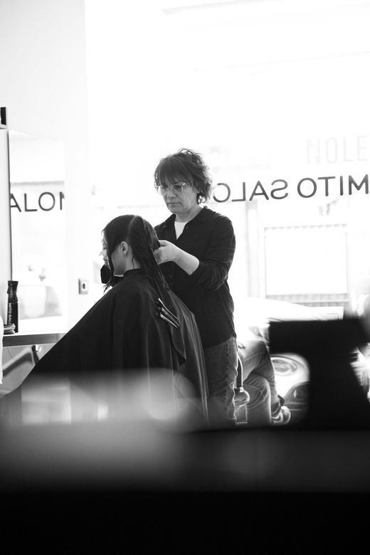 Un parrucchiere taglia i capelli scuri di una cliente in un salone, con specchi e finestre luminose alle sue spalle.