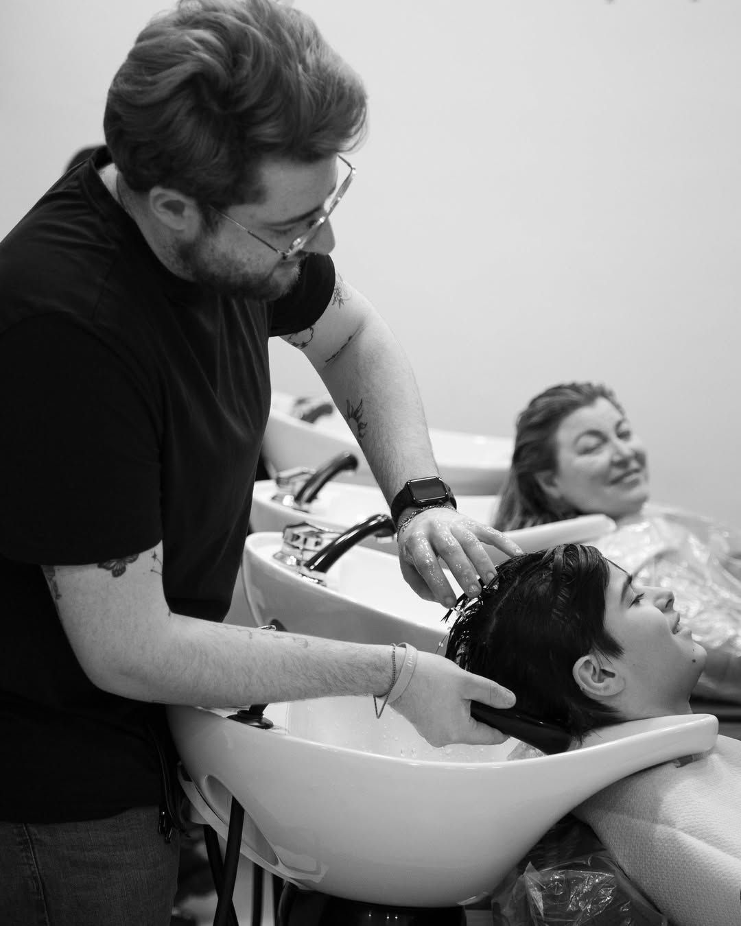 Un parrucchiere lava i capelli a una cliente nel lavandino del salone, mentre un'altra cliente si rilassa nelle vicinanze.