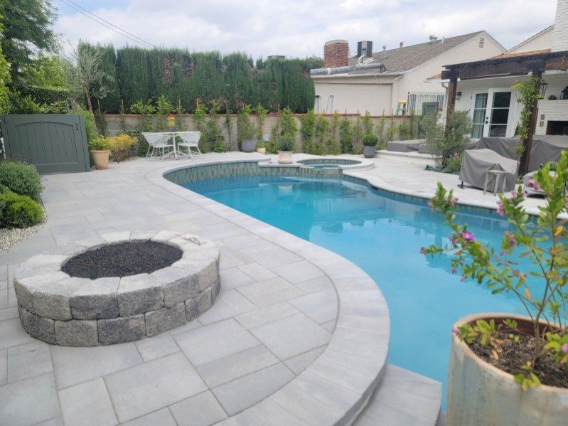 A backyard patio with a stone fire pit, a curved swimming pool, and lounge chairs on a paved deck under a cloudy sky.