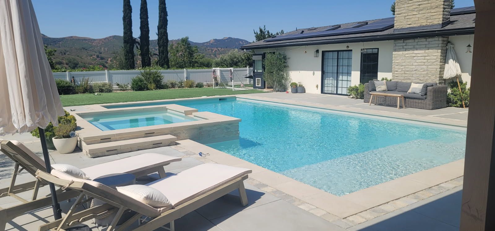 A bright, sunny backyard featuring a swimming pool, attached hot tub, lounge chairs, and a patio area by a house.