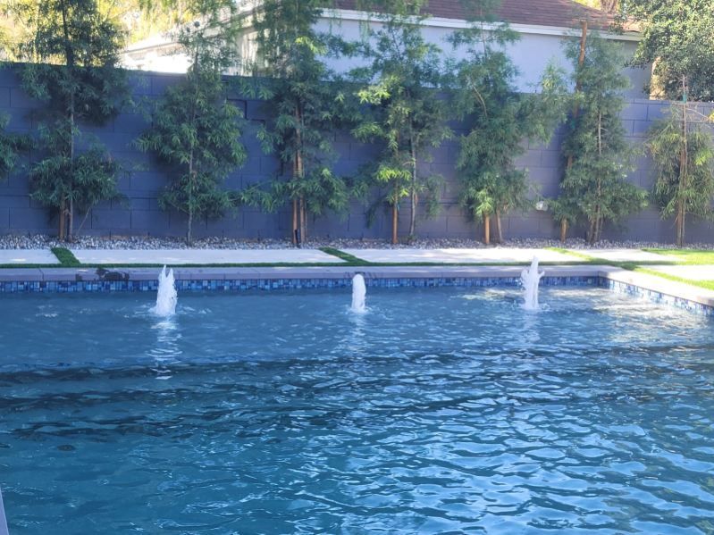 A blue rectangular swimming pool with three vertical water jets spraying upwards, set against a dark wall and tall trees.