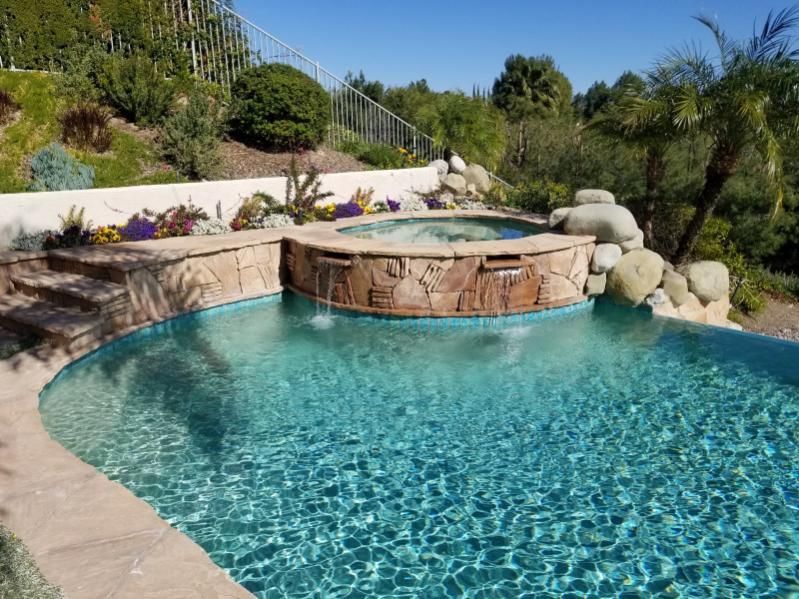 A swimming pool with a raised stone spa and two water features spilling into the clear, blue water on a sunny day.