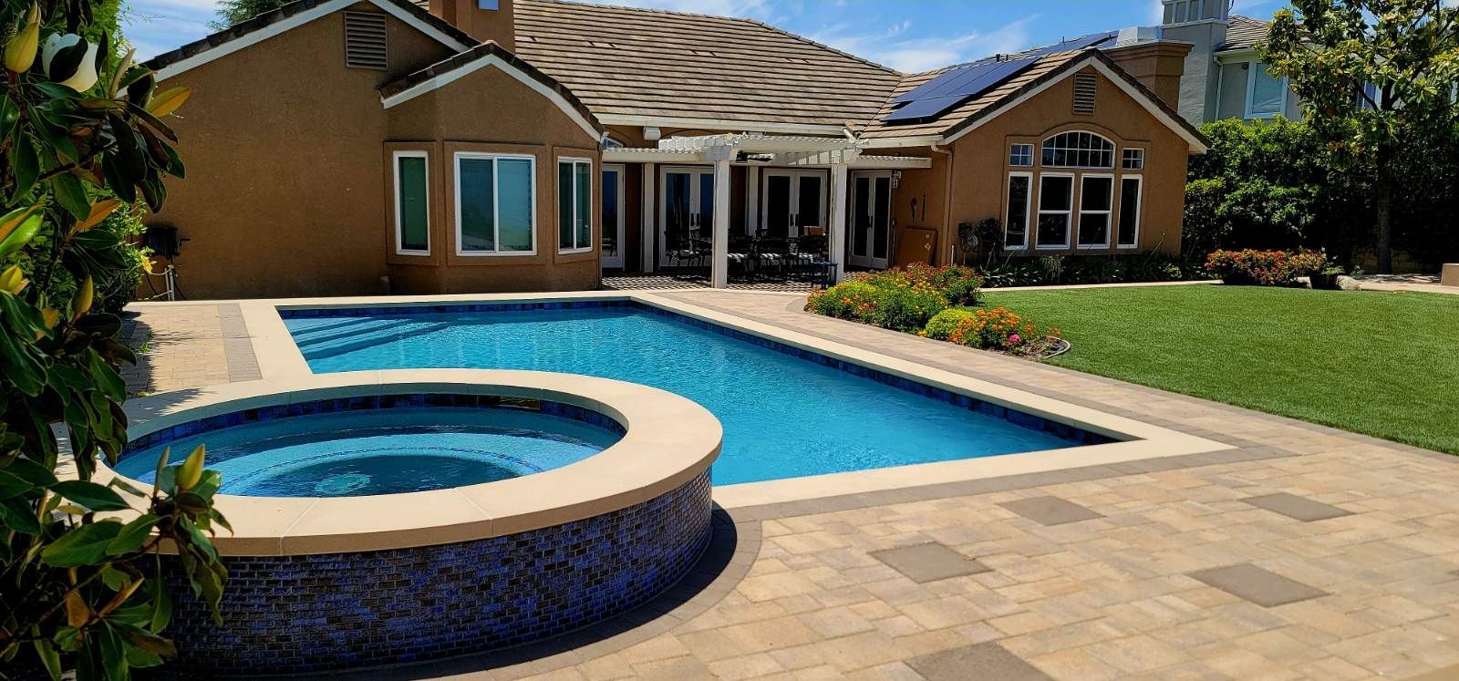A backyard featuring a swimming pool, attached hot tub, paver patio, and lawn next to a tan, multi-roofed house.