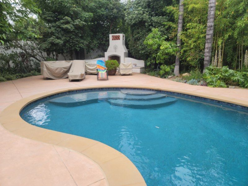 A kidney-shaped swimming pool with blue water and steps, surrounded by a patio, outdoor furniture, and lush green trees.