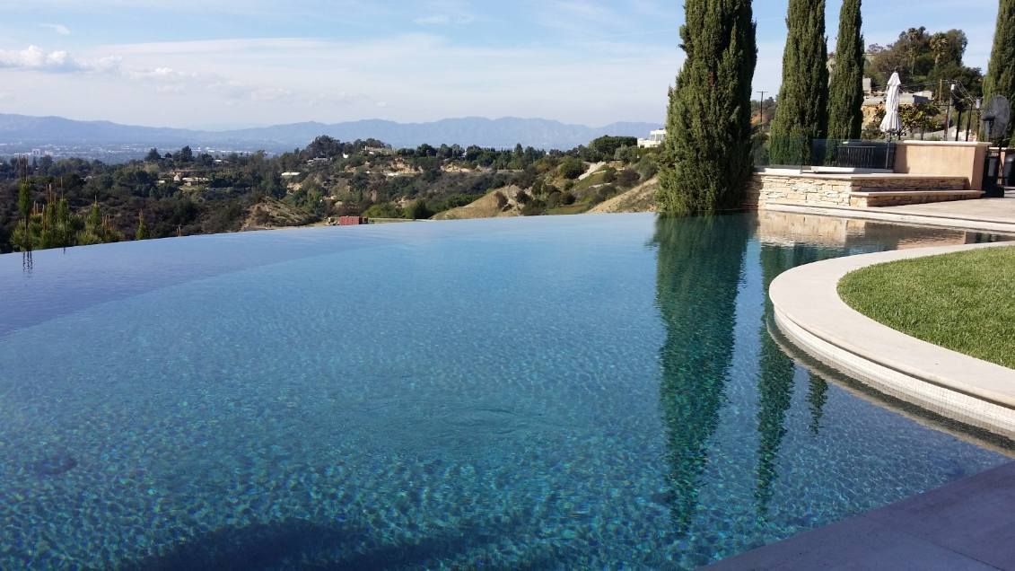 An infinity pool overlooks a scenic valley with rolling hills, tall cypress trees, and a clear blue sky.
