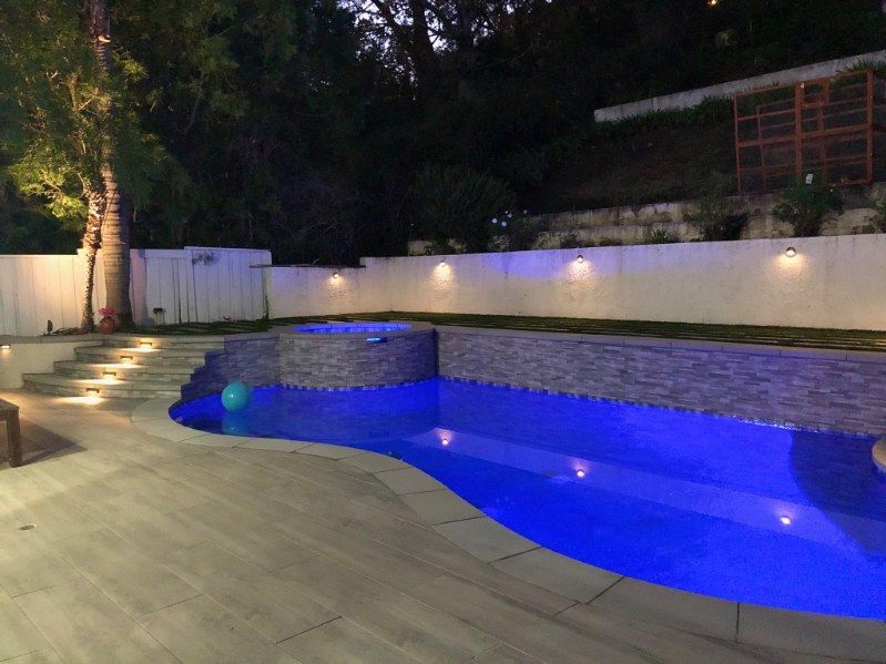A blue-lit backyard swimming pool with a raised spa, stone-textured retaining walls, and tiered steps at night.
