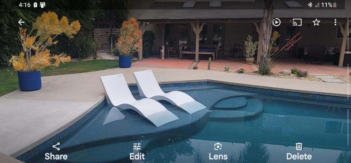 Two white lounge chairs partially submerged in a backyard swimming pool next to a patio with potted plants.