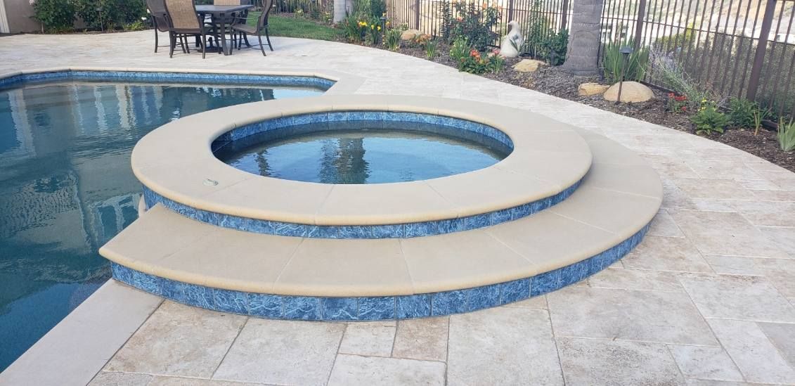 A raised, circular spa with tan stone coping and blue tile accents sits beside a swimming pool on a stone patio.