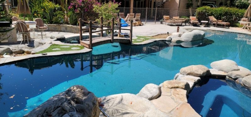 A backyard swimming pool with a wooden footbridge, rocky features, and lounge chairs on the surrounding patio.