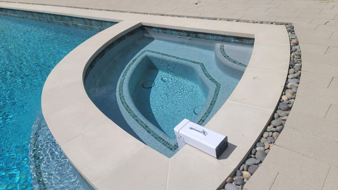 A rectangular device sits on the light stone edge of a blue tiled spa, positioned next to a swimming pool.