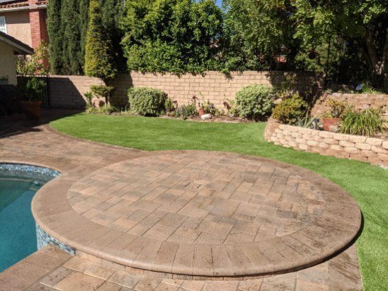 A circular stone patio sits next to a pool, overlooking a manicured lawn and a landscaped stone wall in a backyard.