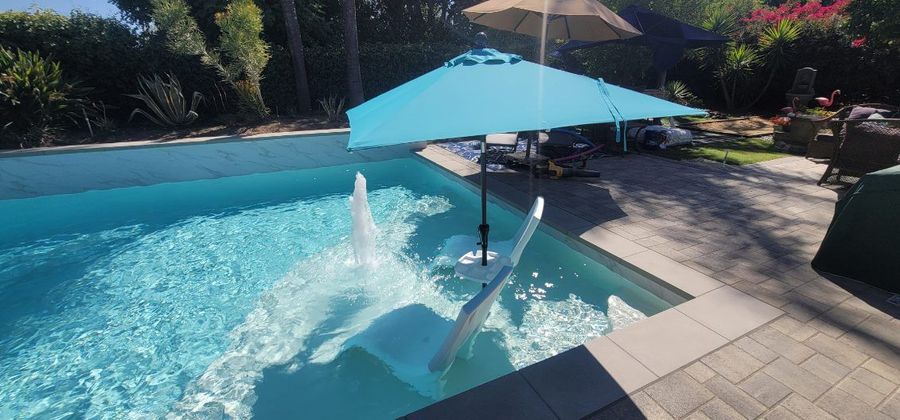 A bright blue pool umbrella stands over a white lounge chair in a sunny swimming pool with a small water fountain.