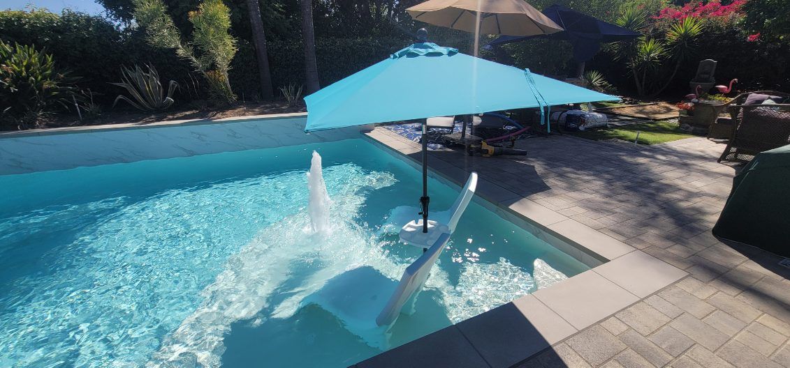A bright blue umbrella shades a white tanning ledge chair in a pool, with a water fountain spraying nearby.
