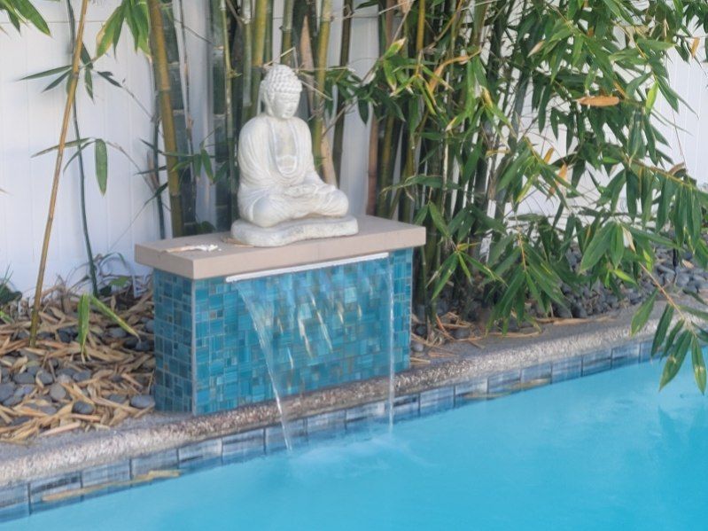 A stone Buddha statue sits on a blue tiled pedestal, with a water feature flowing into a swimming pool beside bamboo.