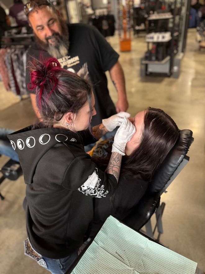 A woman is getting a tattoo on her eyebrow while a man watches.